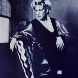 Black-and-white portrait of a glamorous woman in a 1950s-style outfit, leaning on a chair in a studio setting with dramatic lighting.