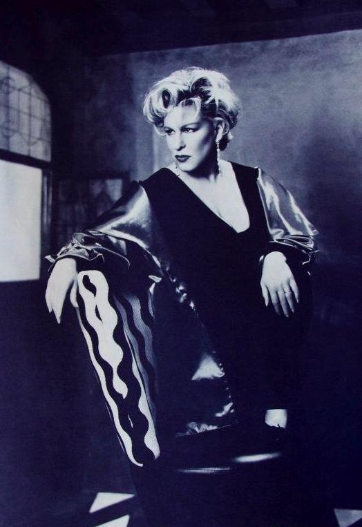 Black-and-white portrait of a glamorous woman in a 1950s-style outfit, leaning on a chair in a studio setting with dramatic lighting.