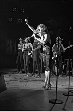 Black-and-white photo of a female lead singer at center stage with a microphone, backed by four backup singers and a guitarist in the background, performing live.