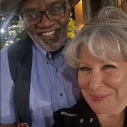 Fab % Freddie and Bette Midler posing for a selfie at night in a city, man wearing a tan fedora, glasses, blue shirt and vest beside a woman with gray hair in a black jacket.