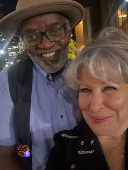 Fab % Freddie and Bette Midler posing for a selfie at night in a city, man wearing a tan fedora, glasses, blue shirt and vest beside a woman with gray hair in a black jacket.