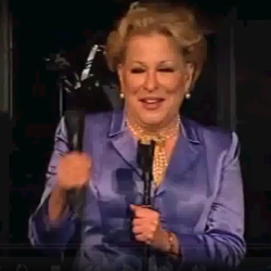 Woman in a purple suit on stage, holding a microphone and an award statue, smiling.