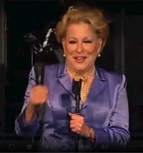 Woman in a purple suit on stage, holding a microphone and an award statue, smiling.