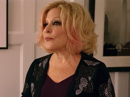 Portrait of a woman with blonde hair with pink tips, wearing a black embroidered jacket, standing indoors beside framed artwork.