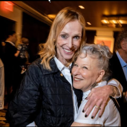 2026-06-02_17-31-55 - bootleg betty Bette Midler & Edie Baskin embrace at an indoor social event; one wears a dark quilted jacket, the other a white top, with warm background lighting.