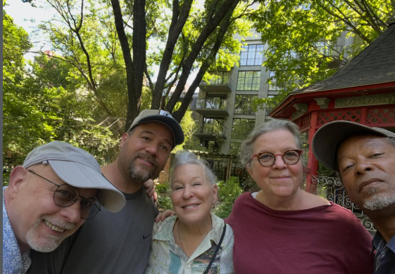 Five adults pose for a selfie in a sunny park, with green trees and a red gazebo in the background.