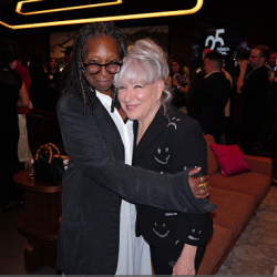 Bette Midler & Whoopi hug and smile for the camera at a social event, with other attendees and lounge seating in a dimly lit venue behind them.