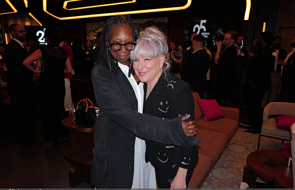 Bette Midler & Whoopi hug and smile for the camera at a social event, with other attendees and lounge seating in a dimly lit venue behind them.