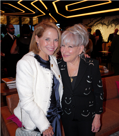 Two women pose together and smile at a social event, one in a white jacket and navy dress, the other in a black suit.