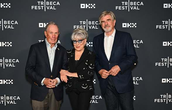 Three people posing on a red carpet at Tribeca Festival, two men in dark blazers with a woman in black with sunglasses in the center.
