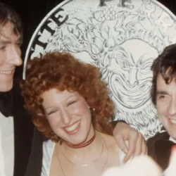 Three people in tuxedos posing and smiling at a formal event, with a large decorative coin backdrop behind them.
