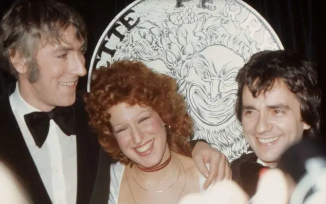 Three people in tuxedos posing and smiling at a formal event, with a large decorative coin backdrop behind them.