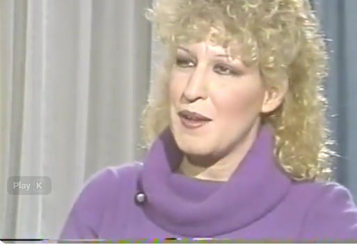 2026-06-09_15-30-00 - bootleg betty Bette Midler with curly blonde hair in a purple sweater being interviewed on a television program.