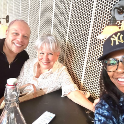 Three friends take a selfie at a table, smiling toward the camera, with a bottle and a phone on the table in front of them against a perforated wall with warm lights.