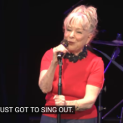 Woman in a red top singing into a microphone on a stage; subtitle reads, "YOU'VE JUST GOT TO SING OUT."