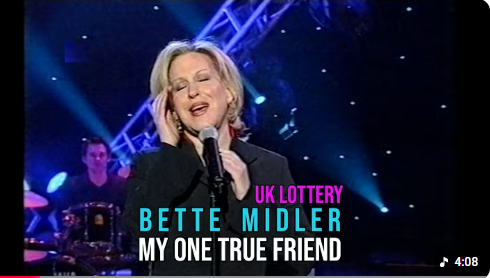 Bette Midler performing 'My One True Friend' on a stage with blue lights for a UK Lottery promo.
