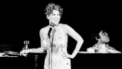 Black-and-white photo of a female singer in a sequined gown at a microphone on stage, with a pianist behind her at a piano.