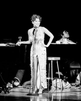 Black-and-white photo of a female singer in a sequined gown at a microphone on stage, with a pianist behind her at a piano.