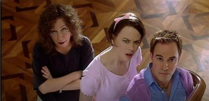 Three adults—two women and a man—look up toward the camera from above, standing on a patterned wooden floor.