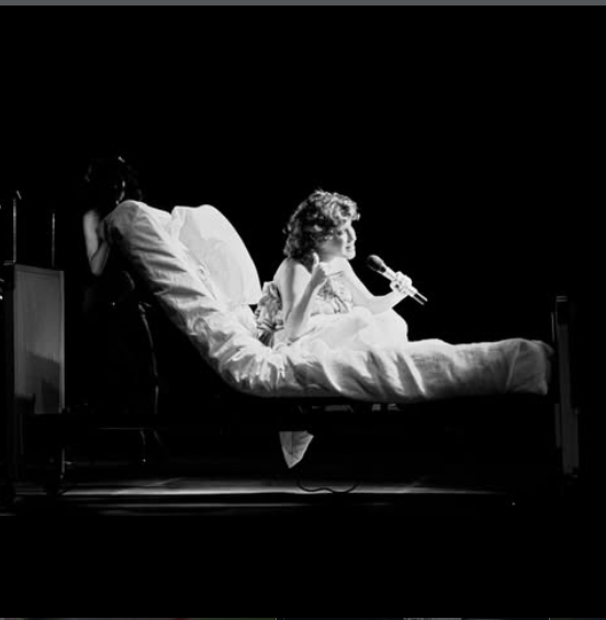 Black-and-white scene of a patient in a hospital bed singing into a microphone with a guitarist seated nearby under dim stage lighting.