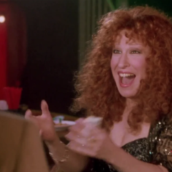 Woman with curly red hair laughing with hands raised at a dimly lit restaurant table.