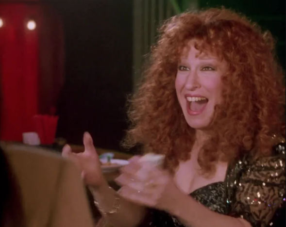 Woman with curly red hair laughing with hands raised at a dimly lit restaurant table.