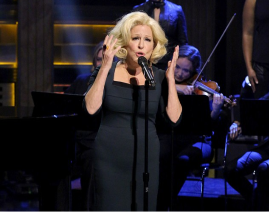 Female singer in a black dress performing on stage with an orchestra in the background, microphone in hand and stage lights overhead.