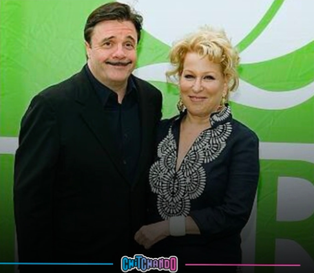 Two people posing and smiling in front of a bright green abstract backdrop; man in a black suit and woman in a black embroidered blouse.