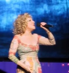Female singer with curly blonde hair performing on stage, holding a microphone in a pastel dress with pink and green tones.
