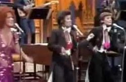Four vocalists perform on a stage with a backing band; a woman in a pink sequined dress sings beside three men in dark suits with pink accents.