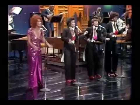 Four vocalists perform on a stage with a backing band; a woman in a pink sequined dress sings beside three men in dark suits with pink accents.