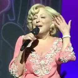 Female singer in a pink embroidered dress sits cross-legged on stage, microphone in hand, performing with a purple-lit backdrop.