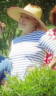Woman wearing a wide-brimmed straw hat and blue-and-white striped shirt, smiling outdoors among greenery.