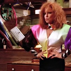 Person with bright, orange hair and a bold multicolor 1980s-style jacket stands in a cluttered workshop, hands on hips, holding a large prop object over their shoulder.
