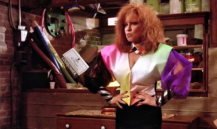 Person with bright, orange hair and a bold multicolor 1980s-style jacket stands in a cluttered workshop, hands on hips, holding a large prop object over their shoulder.