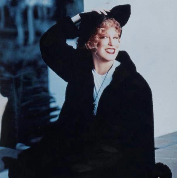 Smiling blonde woman in a black coat and wide-brimmed hat, one hand touching her hat outdoors.