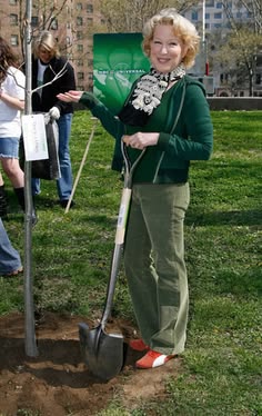 Smiling woman in a green cardigan and scarf gestures with her hand in a park, with a green banner in the background.