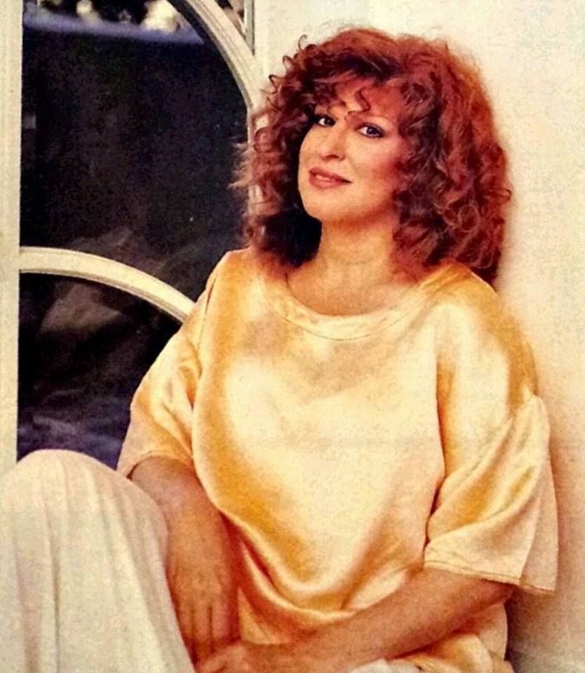 Woman with curly red hair sits against a light wall, wearing a loose peach satin shirt and looking at the camera.