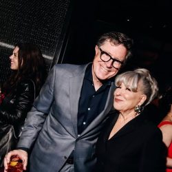 Smiling couple at a party: man in a gray suit with glasses next to a woman with gray hair, both posing together; the man holds a drink in his hand.