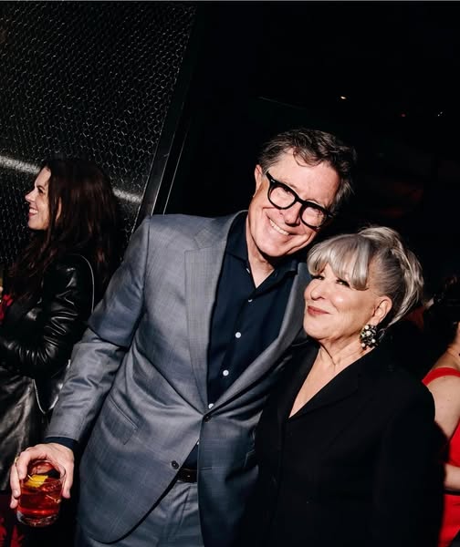 Smiling couple at a party: man in a gray suit with glasses next to a woman with gray hair, both posing together; the man holds a drink in his hand.