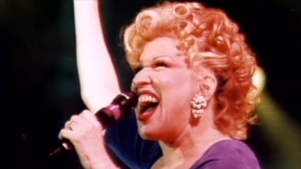 Female singer with blond curls performing on stage, singing into a microphone, wearing a purple outfit and large earrings.