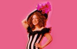 Person in a floor-length black-and-white striped gown with a feathered train, posing with hands on hips against a pink backdrop and pink floral headpiece on the head.