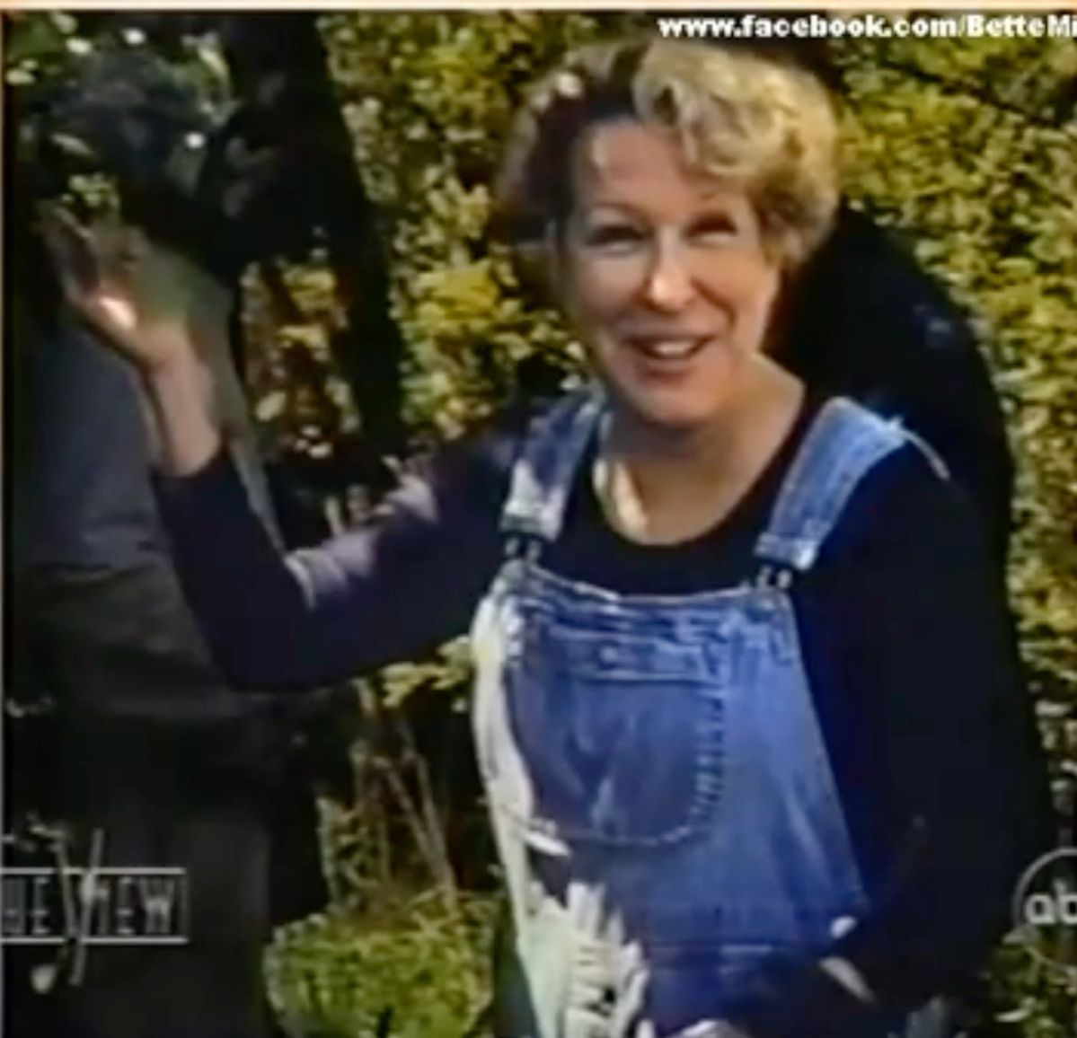 Woman wearing blue denim overalls and a black shirt, smiling and waving outdoors among trees and greenery.
