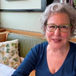 Smiling Bette Midler with gray hair and green glasses sits at a cafe table by a window.