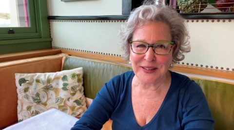 Smiling Bette Midler with gray hair and green glasses sits at a cafe table by a window.