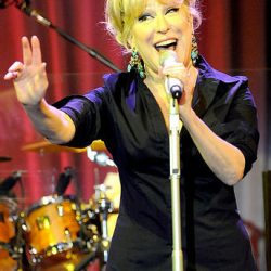 Bette Midler performing on stage, microphone in hand, hand raised, with red curtain and drum set in the background.