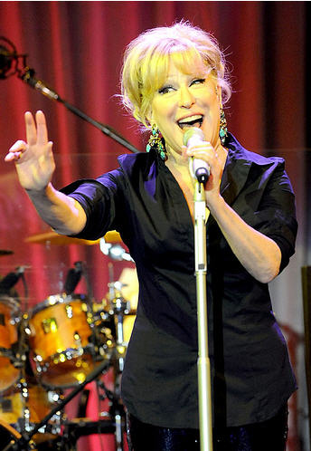 Bette Midler performing on stage, microphone in hand, hand raised, with red curtain and drum set in the background.
