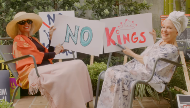 Two women sit outdoors in hats and robes, smiling as they hold signs that read 'NO KINGS' with a small crown illustration.