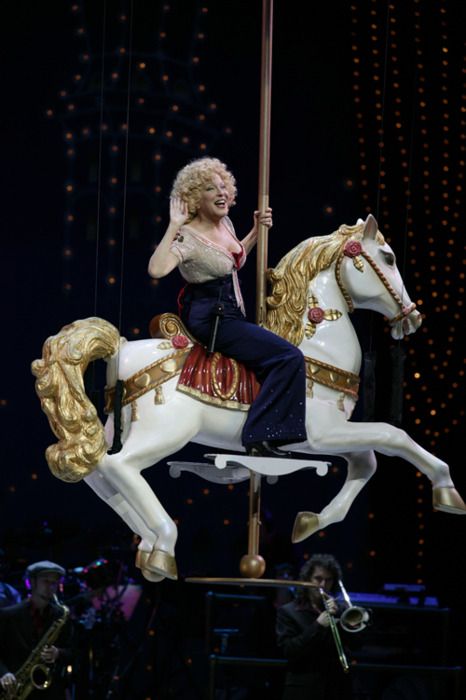 B8967a64b572e0335a1348eb54939f01 1 - bootleg betty Female performer rides a white and gold carousel horse on stage, holding a pole as a band plays in the background.