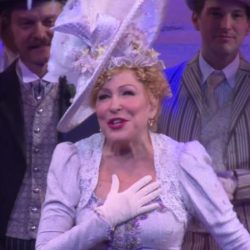 Bette Midler in a Victorian-style lavender dress with white gloves, a decorative hat, and lace, touching her chest on stage.